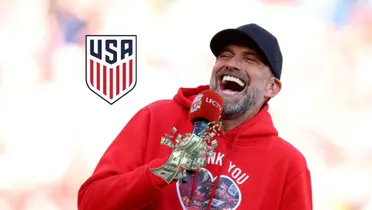 Jurgen Klopp laughs while he wears a special Liverpool sweater while the US National team badge is next to him and money is below him. (Source: The Irish Independent)