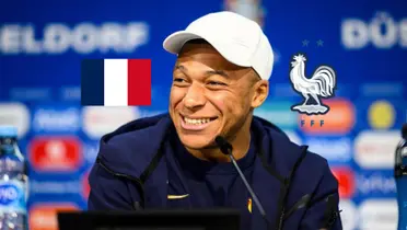 Kylian Mbappé smiles while he talks in a press conference as the French flag and the French national team badge is next to him. (Source: KM 10 Zone)